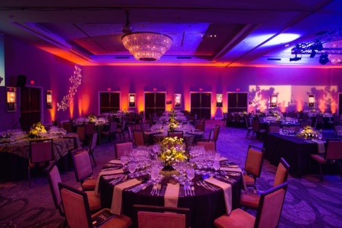 A ballroom with multiple round tables set for a gala, the room washed in shades of pink, blue, and red.