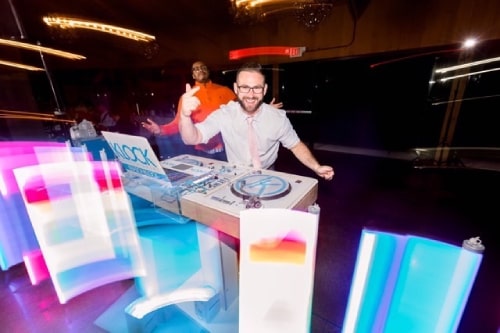 Jason Klock smiling at the camera energetically, leaning over his DJ equipment